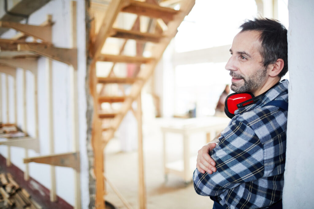 Zufriedener, verträumter, gutaussehender Arbeiter mit Gehörschutz lehnt an der Wand auf der Baustelle und schaut sich um, während er darüber nachdenkt, wie man das Haus renovieren kann