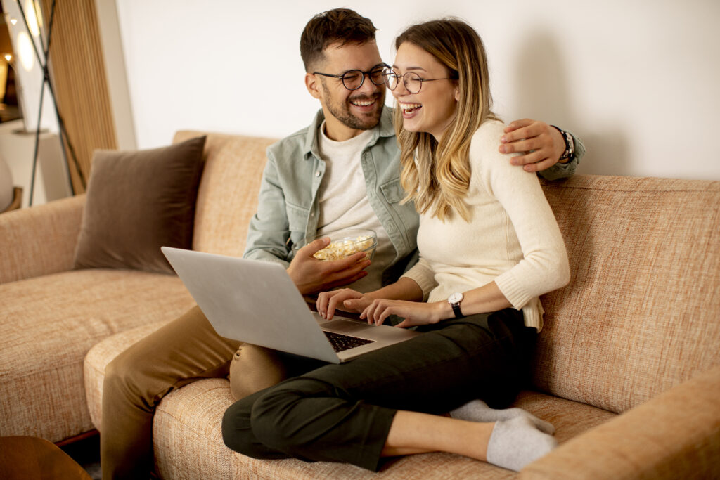 Ein Junges Paar benutzt gemeinsam einen Laptop, während es zu Hause auf dem Sofa sitzt