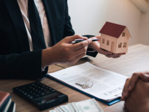 Ein Mensch mit einem Kugelschreiber in der Hand und einem kleinen Haus an einem Schreibtisch, der jemandem eine Finanzierung eines Hauses verdeutlicht