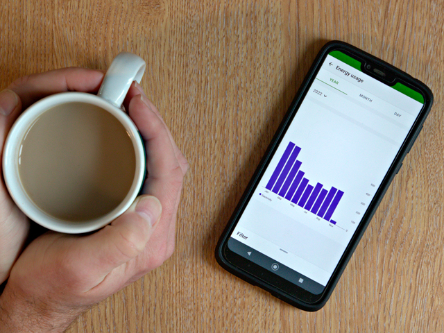 Ein Tisch auf dem ein Smartphone liegt mit einer Statistik über Energie und dazu ein Kaffee der von zwei Händen gehalten wird von oben